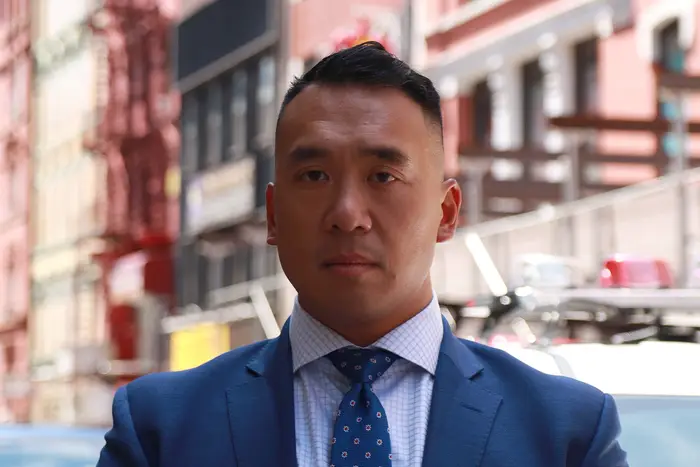 A close-up of Detective Cheung, who is Chinese, standing on a sidewalk. He is wearing a suit and tie.